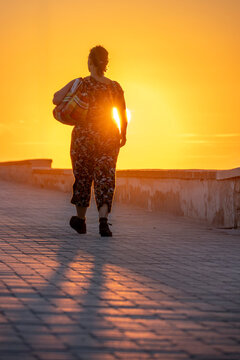 Saint-Aubin-Sur-Mer, France - 07 27 2026: A woman carrying a colorful bag walk along the jetty with the sun at golden hour