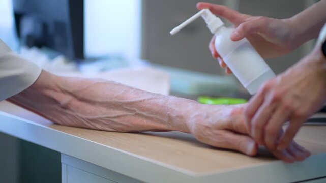 Senior patient receives professional wound disinfection as nurse sprays liquid onto their wrinkled hand in clinic. Elderly individual feels vulnerable yet calm during this sterile treatment process