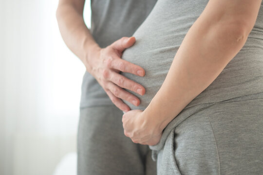 Pregnant woman alone cradling belly with interlaced fingers in serene solo portrait. Expecting mother embracing motherhood in minimal studio with white background showing protective anticipation.