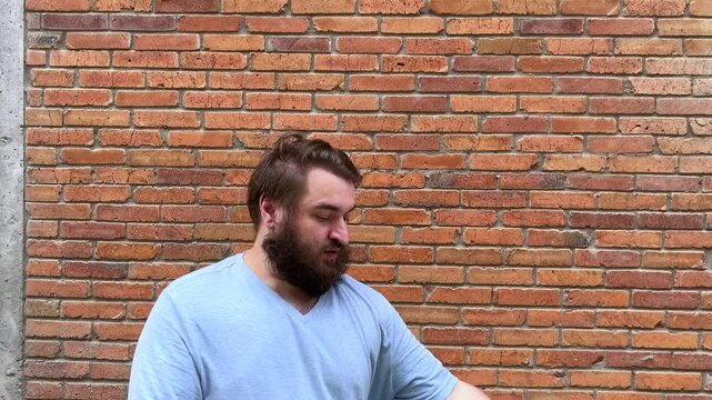4K video. Young adult man with beard stands near house brick wall, picks up and holds blue bundle of soft foam rubber for sofa mattress. Concept of furniture repair, delivery, dream, sleep
