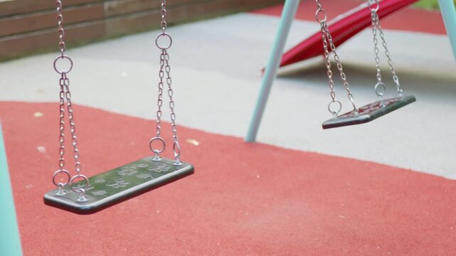 Empty playground swings gently sway in the breeze, with red rubber flooring and a colorful slide visible in the background, creating a serene outdoor play area