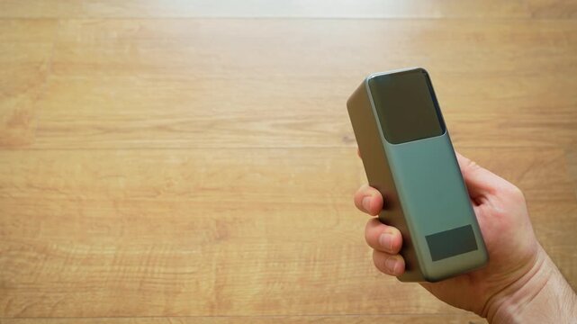 Right hand holding power bank portable charger close up with copy space on left over wooden background. Clean minimal composition focusing on modern external battery device in everyday use. Framing