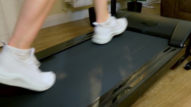 Unrecognizable runner slowing stride on treadmill and stopping after indoor cardio workout. White sport sneakers transitioning from fast running to halt on moving belt in home fitness space. Cardio