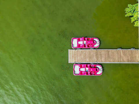 defaultTop down aerial view of two bright pink pedal boats moored at a wooden jetty on a green lake.