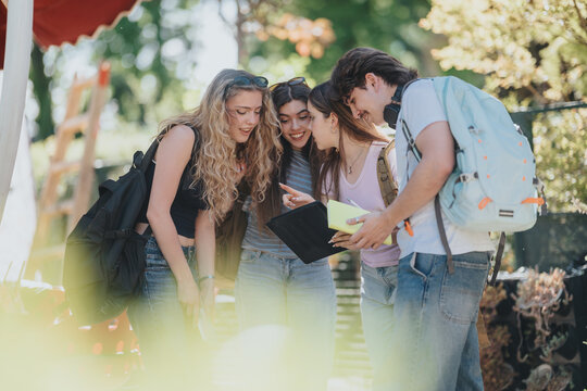 Four teenage students stand outdoors on a sunny campus viewing a tablet and sharing notes. A group of friends collaborate and laugh while checking information during a school break.