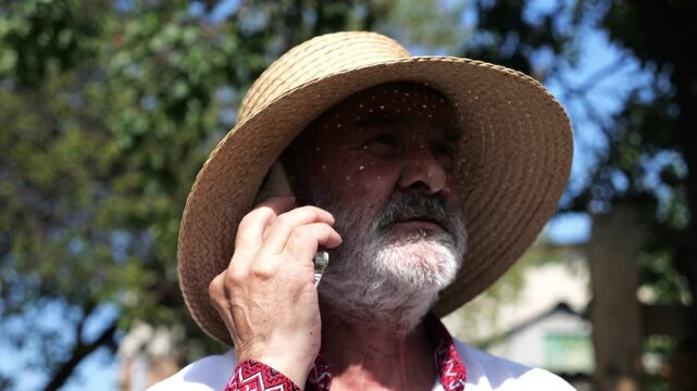 Older ukrainian man wearing in vyshyvanka talking on mobile phone at summer garden. Senior male in straw hat using cell phone outdoor. Concept of connection between tradition and modern life