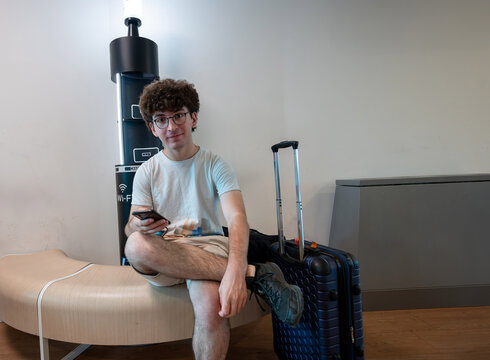 Young man sitting on a modern bench, holding a smartphone, and smiling at the camera while waiting for his flight with luggage at an airport lounge