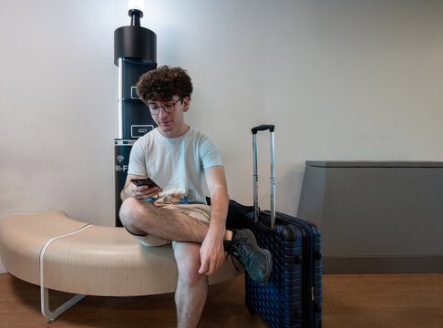 Young man sitting on a modern bench, checking his smartphone next to a charging station with wi fi signal, and his blue suitcase, waiting for travel in an airport lounge