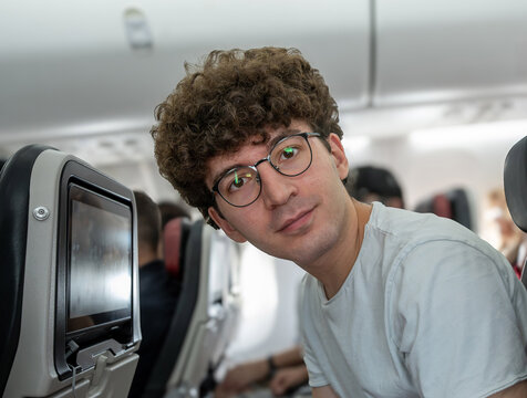 Young man with curly hair and glasses looking directly at the camera while traveling in the passenger cabin of an airplane, offering a friendly expression during his flight journey