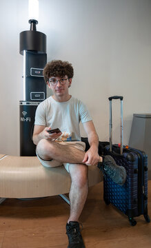 Young man sitting on a bench in an airport lounge, waiting for travel, holding a smartphone, and connected to a public charging station with luggage beside him, accessing wi fi