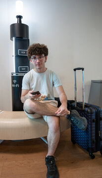 Young man with curly hair and glasses sitting by a charging station in an airport lounge, checking his smartphone while waiting with luggage