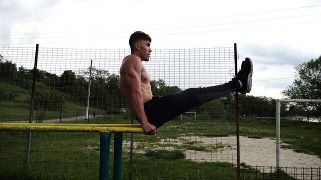 Strong man trains abdominal muscles raising legs on parallel bars at sports ground. Young shirtless sportsman exercising at nature. Sporty athlete working out at nature. Concept of sportive lifestyle