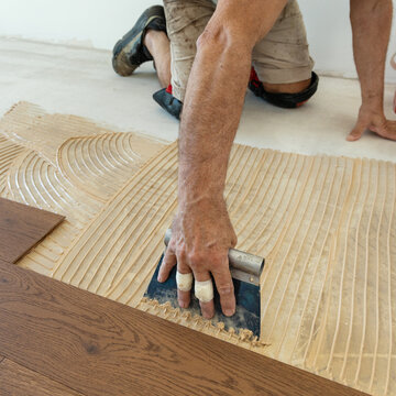 Worker applying adhesive on concrete floor with notched trowel during oakwood parquet installation