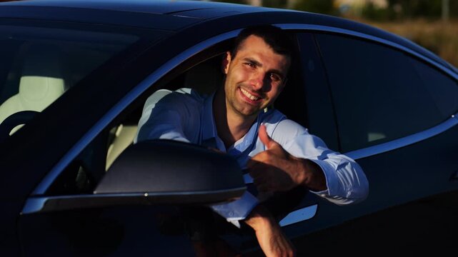 Young smiling entrepreneur leaning out of driver window at modern electric car and looking into camera. Portrait of handsome successful businessman showing thumbs up after bought new electrical auto