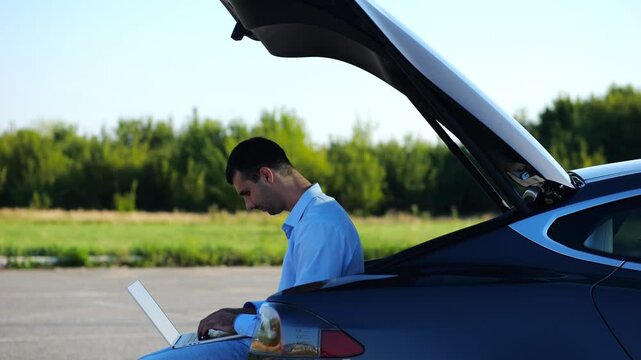 Young handsome entrepreneur works on notebook sitting in electric car trunk. Successful businessman in formal wear using computer at auto. Confident manager typing text on laptop outdoor. Slow mo
