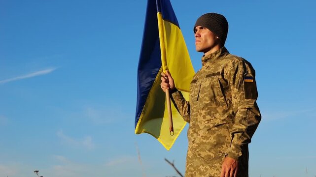 Ukrainian army man stands with raised national banner to peak of hill. Male soldier in military uniform with Ukraine flag as symbol of victory against russian aggression. Invasion resistance concept