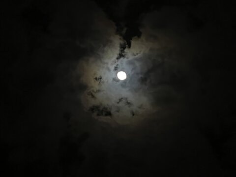 Mystical full moon shining through dark clouds at night, glowing lunar sphere in a dramatic evening sky, ethereal moonlight background for spooky themes