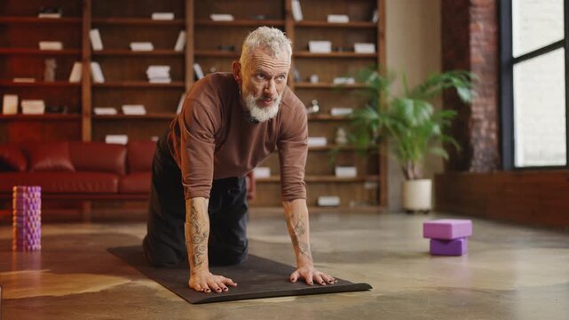 Handsome tattooed man in his 50s performing tricky kitty yoga pose at home