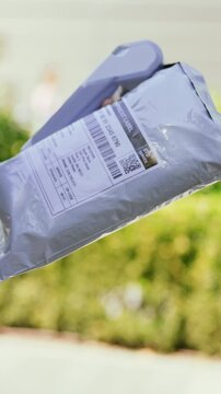 Vertical video. A delivery person stands outside a building and hands over a package to someone. There are plants and grass in the background while the sun is shining in the day.
