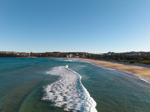 Aerial view of Dee Why Beach with turquoise waves breaking on the sandy shore, coastal vegetation, and urban buildings under a clear blue sky in Dee Why, New South Wales, Australia.