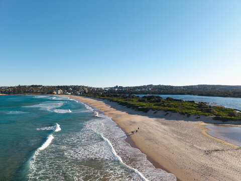 Aerial view of Dee Why Beach with gentle ocean waves rolling onto the sandy shore, coastal vegetation, and people walking along the coastline in Dee Why, New South Wales, Australia.
