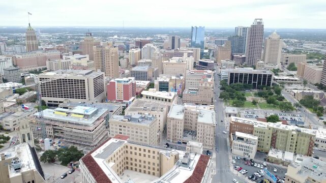 Downtown district Travis Park spans commercial blocks adjacent to historic municipal structures. Vehicular traffic navigates grid streets beneath overcast skies in San Antonio, TX