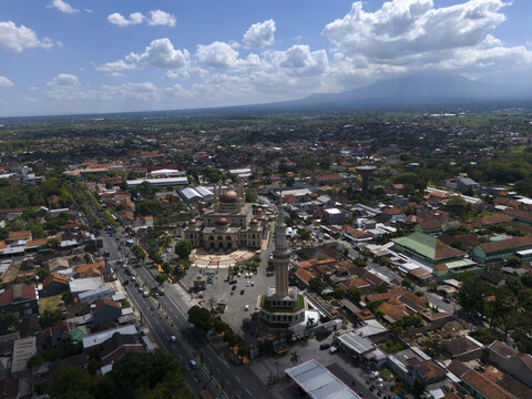 Aerial view of Al Aqsha Great Mosque Klaten surrounded by urban rooftops, roads, and lush greenery under a bright blue sky in Kecamatan Klaten Utara, Jawa Tengah, Indonesia.