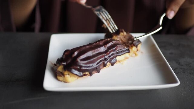 Woman cutting chocolate eclair with fork and knife on white plate against dark background