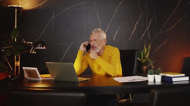 Busy company CEO in his 50s talking on phone while working on a laptop in office