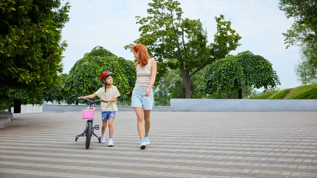 Young girl confidently rides bicycle while mother walks nearby in park. Concept of cycling confidence, positive parenting, relaxed summer leisure.