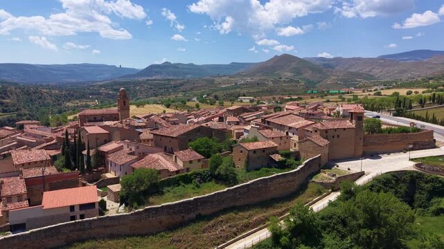 Hilltop walled village of Mirambel, Teruel, Spain, with terracotta rooftops and church tower rising above green terraced farmland and layered mountain ridges under partly cloudy sky