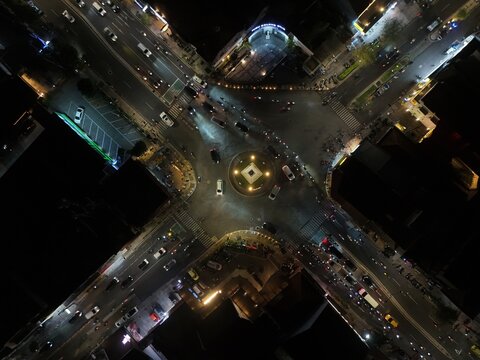 Aerial view of Tugu Yogyakarta Monument illuminated at night with traffic moving through the intersection and surrounding urban buildings in Kecamatan Jetis, Daerah Istimewa Yogya, Indonesia.