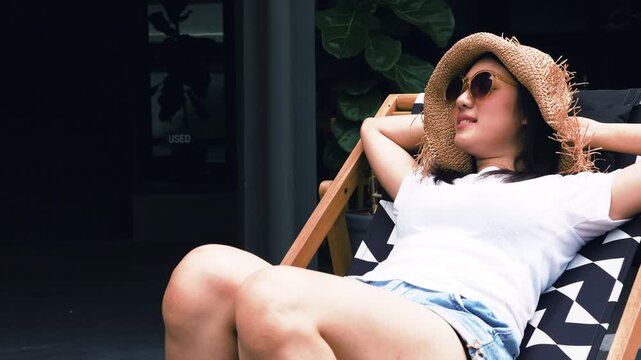 Relaxed young woman wear straw sun hat and sunglasses rest on deck chair outdoors She enjoys peaceful sunny day representing summer leisure, vacation, self care, slow living lifestyle