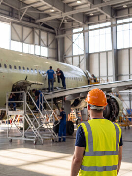 A technician in a highvisibility vest and hard hat overseeing aircraft maintenance in a large hangar facility