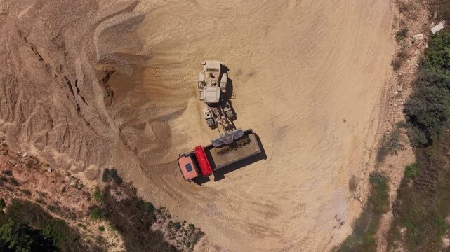 Top-down nadir view of a wheel loader depositing aggregate material into a red dump truck bed at a quarry site, with sweeping tire tracks across pale sandy ground