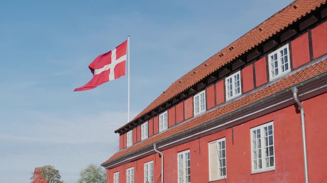 Danish national flag waving above traditional red timber frame building on sunny day with blue sky.