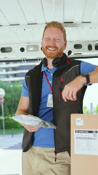 Vertical video of a man stands inside a delivery van with boxes around him. He checks a package while getting ready to deliver items. This takes place outside in a city area on a sunny day.