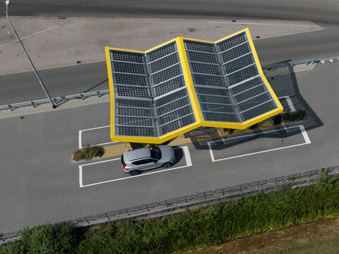 Aerial view of a modern solar-powered electric vehicle charging station with a distinctive yellow-framed zigzag roof and a silver SUV parked underneath in the Netherlands.