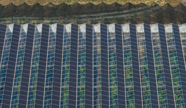 Aerial view of agrivoltaic greenhouse roofs with photovoltaic panels forming geometric patterns and dramatic shadow lines in Saulce-sur-Rhone, Auvergne-Rhone-Alpes, France.