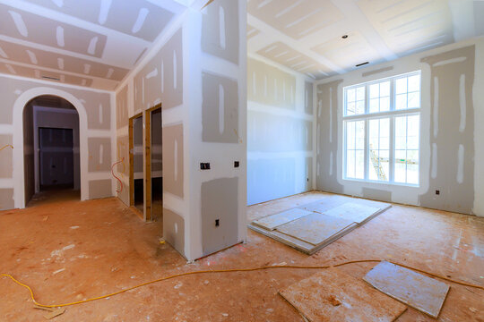 Workers are preparing new home interior with drywall, bare floors in room with large windows.