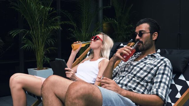 Happy young couple relaxing on deck chairs outdoors, wearing sunglasses, drinking colorful beverages from glass bottles and using digital tablet together. Joyful man and blonde woman enjoying summer v
