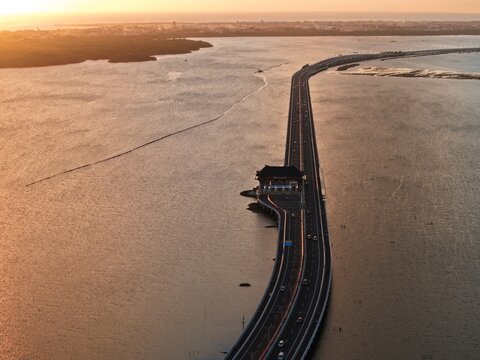 Aerial view of the Bali Mandara Toll Road and its toll plaza stretching across the water during a golden sunset in Kabupaten Badung, Bali, Indonesia.