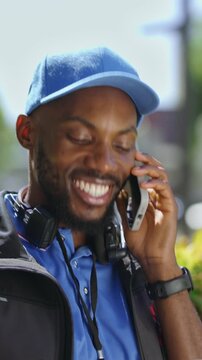 Vertical video of a delivery worker talks on the phone while standing outside. He smiles as he holds a box in his hand. The setting shows greenery and vehicles in the background.