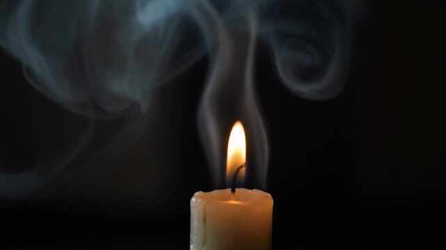 A lit candle flame burning brightly with wisps of white smoke rising against a dark black background in close up.