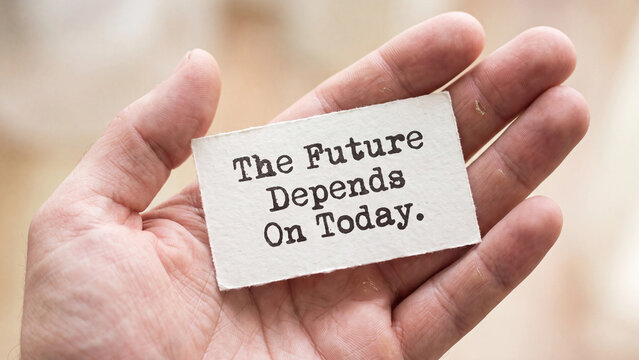 A hand holding a small piece of paper with the text the future depends on today written on it