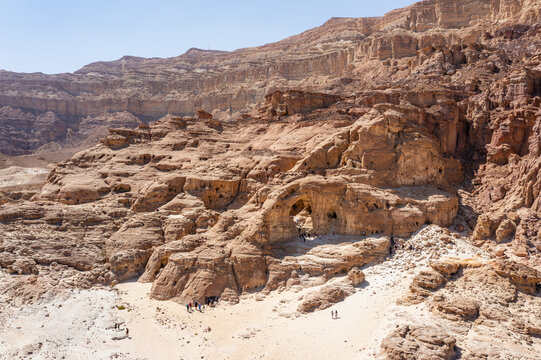 Aerial view of the natural rock arch at Timna Park surrounded by rugged desert cliffs and sandy terrain with visitors exploring the area in Eilat, South District, Israel.