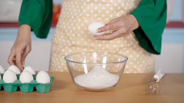Woman's hand breaking a raw egg in a bowl of all-purpose flour - baking concept
. Indian female baker in a printed apron  adding an egg to refined flour / maida bowl kept on a wooden table
