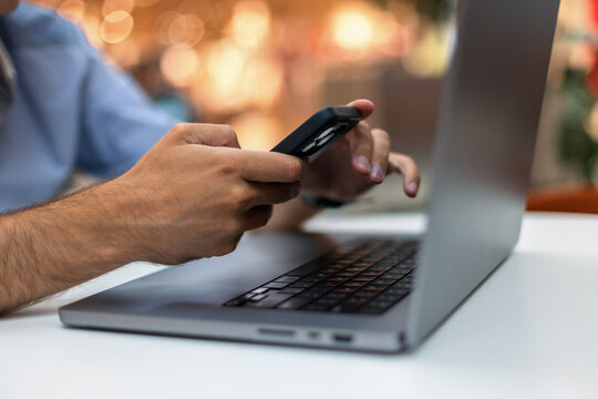 Close-up of a man's hands using a smartphone while working at a laptop on a white desk, with a warm bokeh background. Perfect for concepts of multitasking, remote work, and mobile banking.