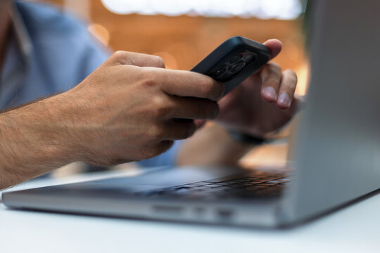 Close-up of a man's hands using a smartphone while working on a laptop computer. Blurred cozy background with warm bokeh lights. Ideal for technology, remote business, and multitasking concepts.