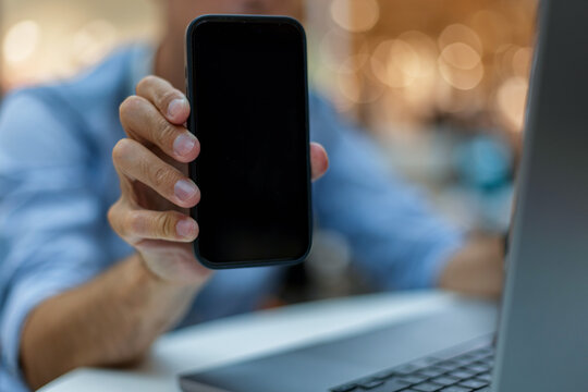 Close-up of a man holding a smartphone with a blank black screen towards the camera while working on a laptop at a desk. Perfect mockup for mobile application or digital business promo.